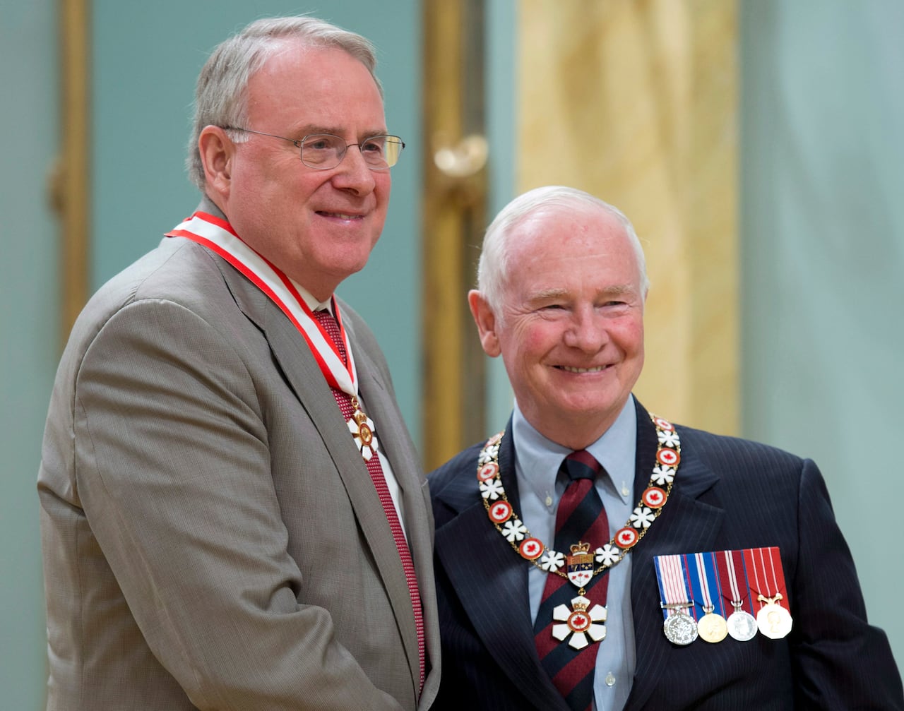 A tall grey-haired man wearing glasses and a grey suit, with a medal hanging around his neck, shakes hands with a white-haired man in a dark suit displaying medals.