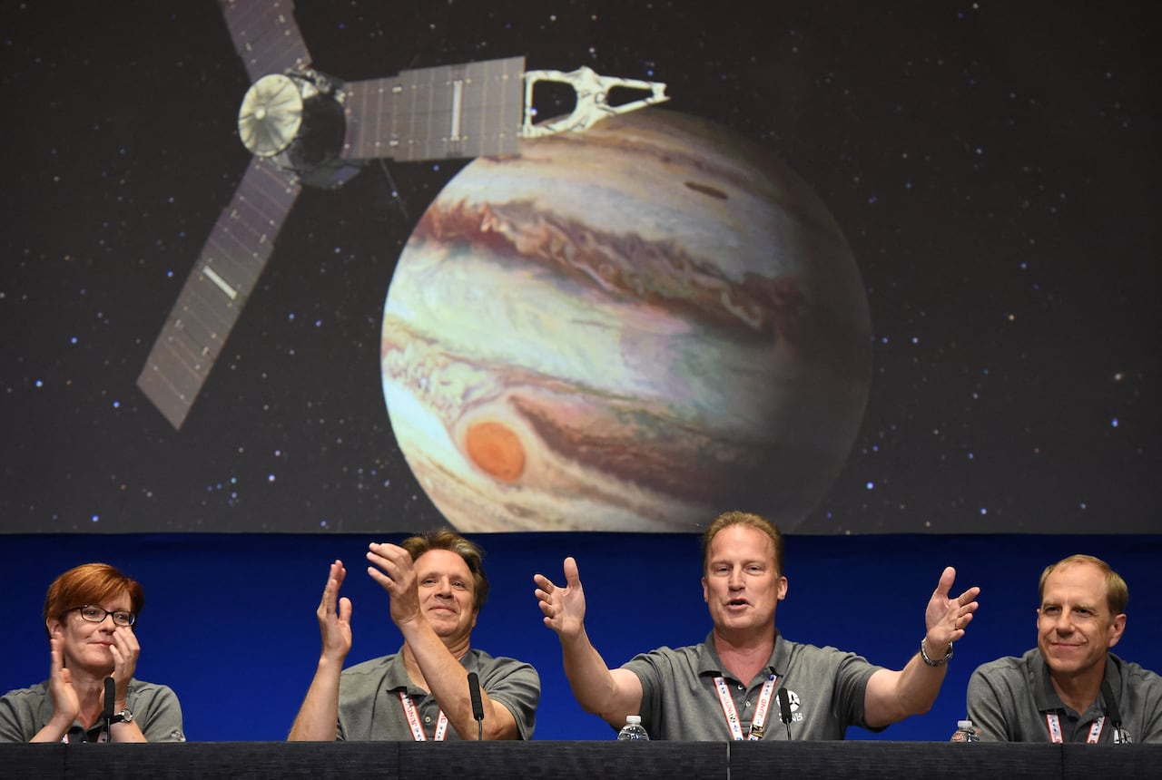Four people presenting at a press conference, clapping and cheering. Behind them is an illustration of a spacecraft and Jupiter.