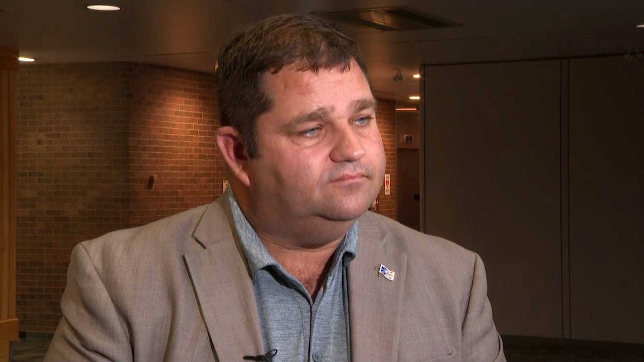 A man in a beige suit jacket and grey shirt stands in the lobby of a large building.