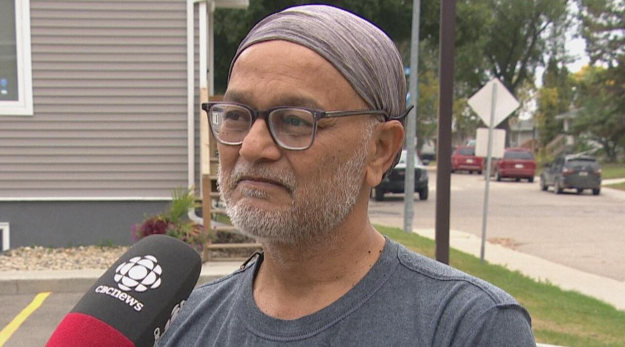A man in a grey shirt and grey head covering, and wearing glasses, speaks into a CBC microphone.