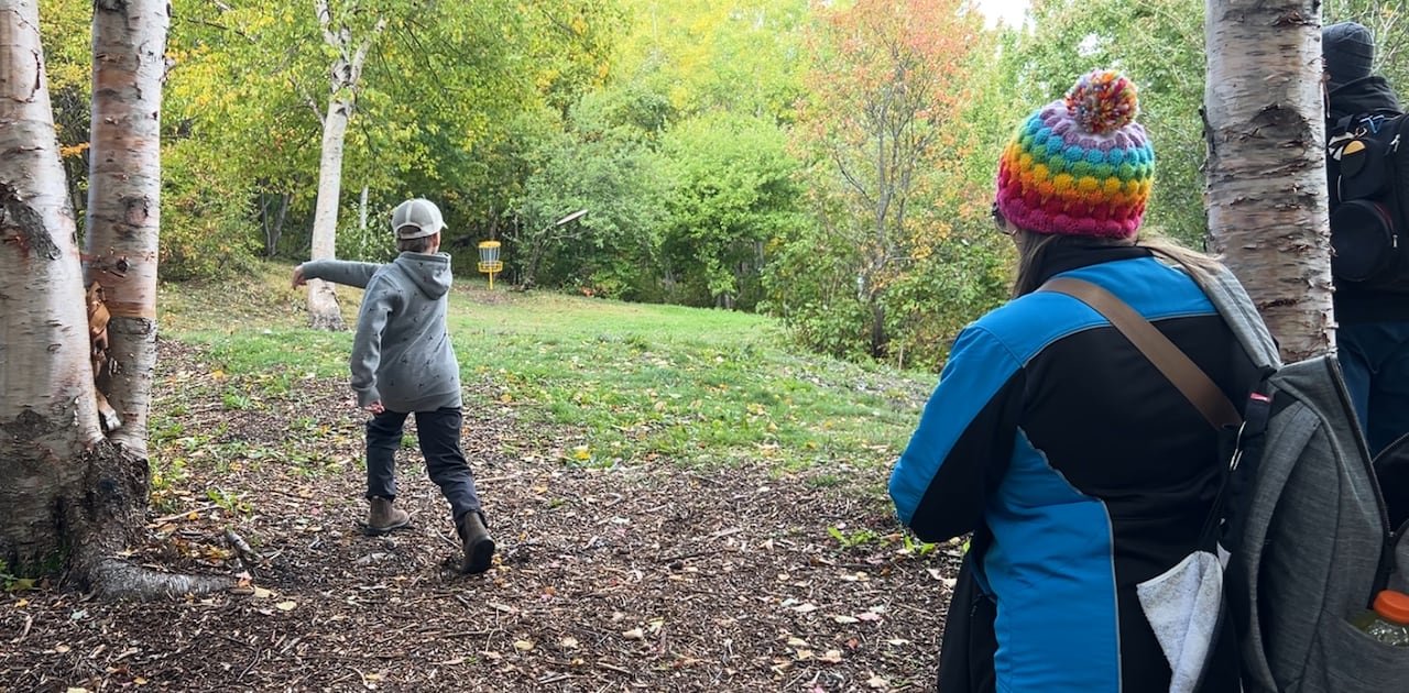 A boy throws a Frisbee on a disc golf course. His parents are watching him.