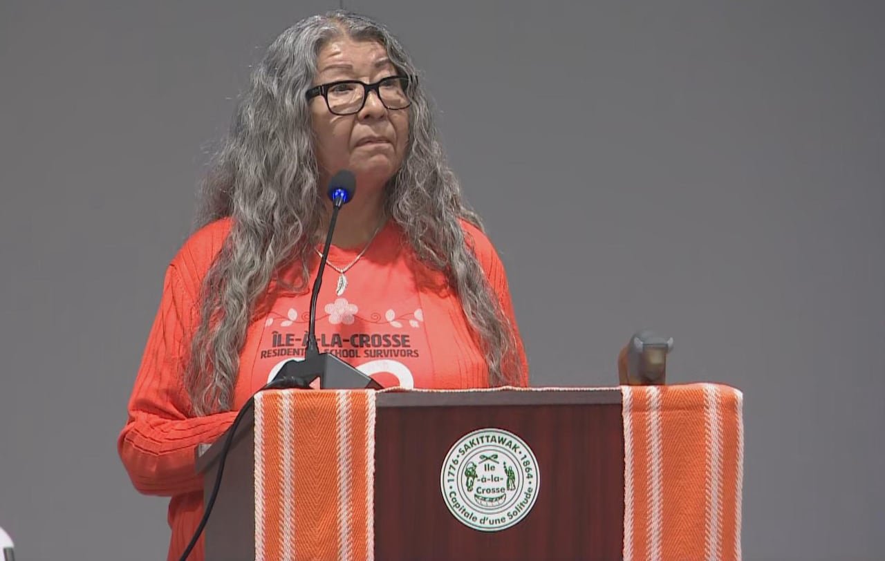 A woman with long grey hair and wearing an orange shirt speaks at a podium.