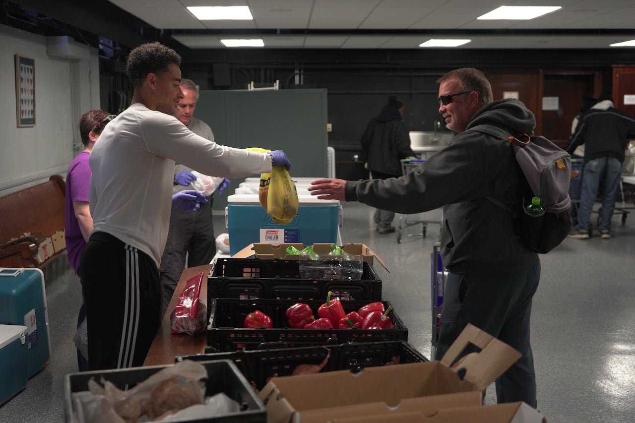 default-140 A young man hands a bag to an older man wearing sunglasses and a coat in what appears to be a transaction. The two people are separated by a table with fruits and vegetables on it.