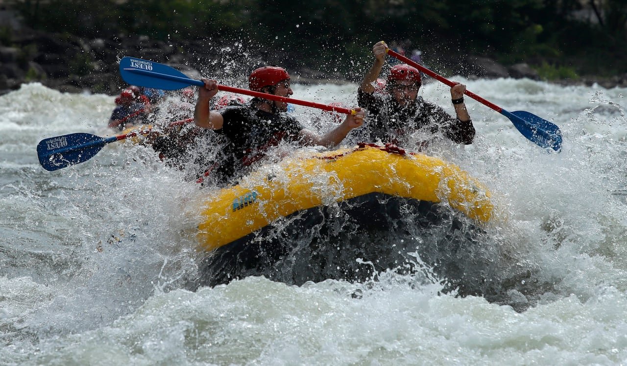 Water splashes against a boatful of people going down some river rapids, as they hold paddles in the air.