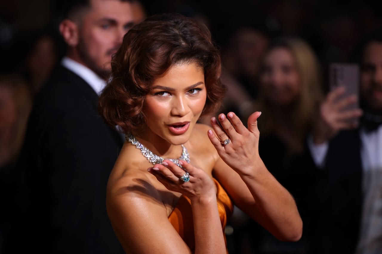 A woman blows a kiss, diamond rings can be seen on the fourth finger of both hands, she is also wearing a large diamond necklace.