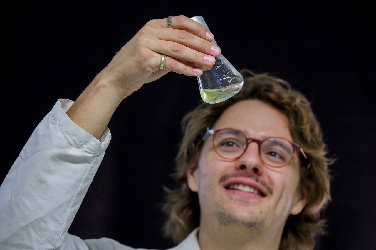 A bespectacled, mustachioed male in a lab coat holds up a beaker with a substance in it.